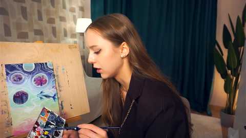 Media: Video of a young woman with long brown hair, wearing a black shirt, painting abstract art on a canvas. She has a small, delicate necklace. Background features a teal curtain, modern wallpaper, and a green plant.