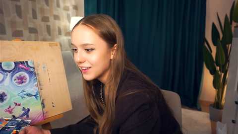 Media: Video of a young Caucasian girl with long, straight brown hair, seated at an easel painting a colorful abstract floral design. She wears a black jacket. The room features teal curtains, a potted plant, and a geometric-patterned wall.