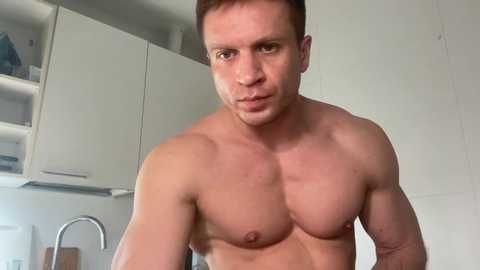 Media: Video of a shirtless, muscular, fair-skinned man with short brown hair and a serious expression in a modern kitchen with white cabinets and a silver faucet.