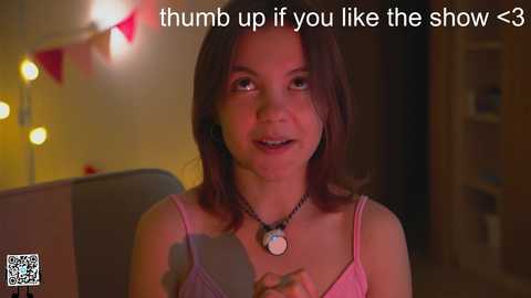 Media: Video of a young woman with shoulder-length dark hair, wearing a pink spaghetti-strap top, in a dimly lit room with a lamp and string lights. Text overlay: \"thumb up if you like the show :)\