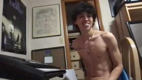 Media: Video of a young, shirtless man with messy hair, smiling, in a cluttered room with posters, shelves, and a wooden chair.