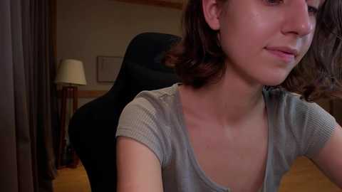 Media: Video of a fair-skinned woman with shoulder-length brown hair, wearing a gray shirt, seated indoors. Background features a black chair, wooden floor, beige walls, and a lamp.