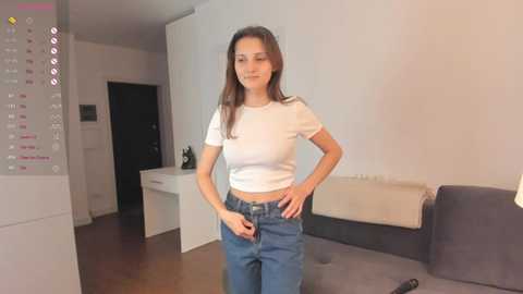 Media: Video of a slender, light-skinned woman with long brown hair, wearing a white crop top and high-waisted jeans, standing in a modern, minimalist living room.