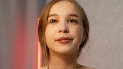 Media: Video of a young, light-skinned woman with straight, shoulder-length light brown hair and fair skin. She has a neutral expression, wearing minimal makeup and a beige top. The background is blurred with soft pink and orange hues.