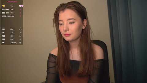 Media: Video of a young Asian woman with straight, long brown hair, wearing a black off-shoulder top, seated indoors against a beige wall.