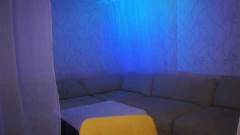 Media: A dimly lit, cozy room with a blue light casting a serene ambiance. A beige sectional sofa with a yellow cushion and a white throw blanket is prominently displayed.