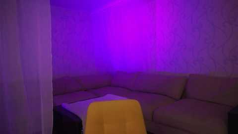 Media: A video of a dimly-lit, cornered living room with purple light. A yellow chair and a black couch with beige cushions are visible, against white wallpaper with subtle patterns.