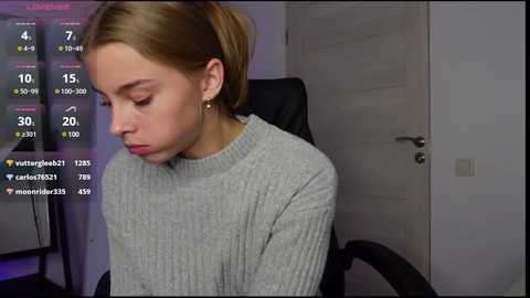 Media: Video of a young, fair-skinned woman with blonde hair tied back, wearing a gray ribbed sweater, sitting in a black office chair. She appears tired, with a blank expression.