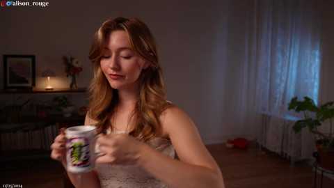 Media: A video of a fair-skinned woman with long, wavy auburn hair, wearing a white lace dress, holding a colorful ceramic mug. The background features a dimly lit room with a fireplace, curtains, and a potted plant.