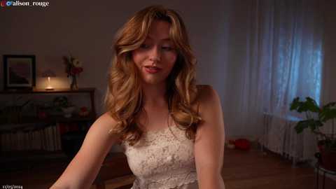 Media: Video of a fair-skinned woman with wavy, shoulder-length blonde hair in a cream lace dress, seated indoors. Background includes a fireplace, potted plants, and sheer curtains.