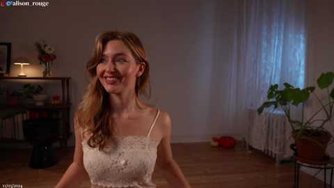 Media: Video of a smiling, light-skinned woman with long, wavy blonde hair in a white lace dress, standing in a dimly lit room with a window, potted plants, and a radiator.