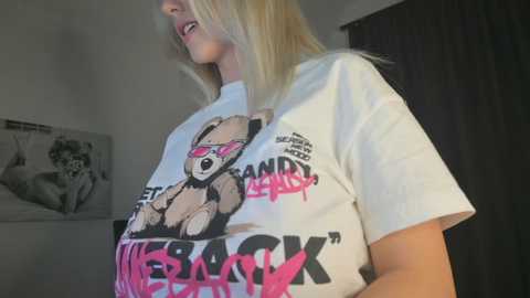 Media: A video of a blonde woman wearing a white t-shirt with a graphic of a teddy bear and \"BACK\" written in graffiti. She's in a dimly lit room with black curtains and a monochrome poster on the wall.