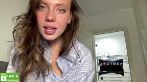 Media: A video of a young woman with wavy brown hair, wearing a striped shirt, smiling and sticking out her tongue, standing in a bedroom with a bed and white walls.