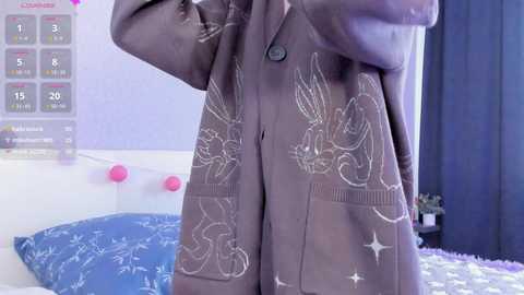 Media: Video of a person wearing a gray, long-sleeve, graphic hoodie with white, hand-drawn illustrations of rabbits and stars. The background features a bed with a blue, star-patterned duvet and pink wall-mounted lights.