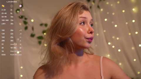 Media: A video of a light-skinned, blonde woman with medium-length hair, wearing a light pink bralette, standing in a softly lit room with fairy lights and greenery in the background.