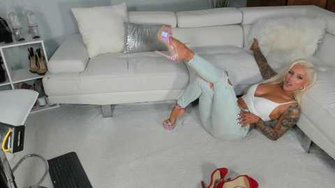 Media: Video of a blonde woman with tattoos lying on a white couch, wearing a white bra and light blue jeans, legs raised, in a modern living room.