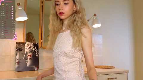 Media: Video of a slender, fair-skinned woman with long, wavy blonde hair wearing a sleeveless, white lace dress. She stands in a modern bathroom with a tiled wall, a mirror, and a \"GQ\" magazine cover.