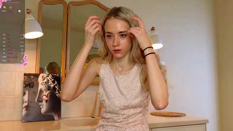 Media: Video of a young, slender, blonde woman with long hair, adjusting her bangs in a bathroom. She wears a sleeveless lace dress, with a mirror and wall calendar behind her.