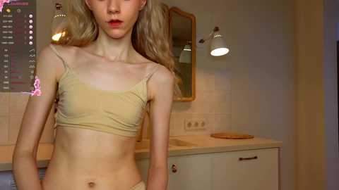 Media: Video of a slender, young, androgynous person with pale skin, wearing a beige bra and pants, standing in a beige-tiled bathroom with a wall-mounted mirror and light fixture.