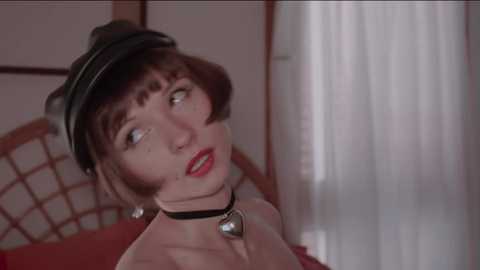 Media: Video of a fair-skinned woman with short brown hair, wearing a black beret, red lipstick, and a black choker with a bell, gazing thoughtfully. Background features a wooden screen and a red curtain.