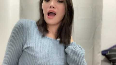 Media: Video of an Asian woman with straight black hair, wearing a light blue knit sweater, mouth open, in a blurred bathroom setting.