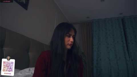 Media: Video of a young woman with long, straight black hair, wearing a red jacket, sitting on a bed in a dimly lit room with green curtains.