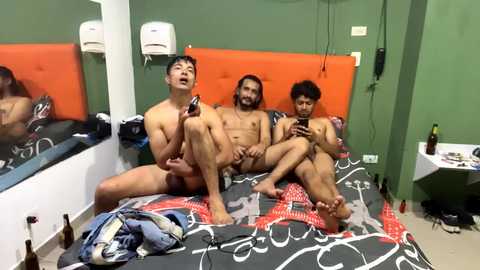 Media: Video of three nude Asian men sitting on a bed, smiling and holding smartphones. Background features green walls, a mirror, orange headboard, and scattered clothes.