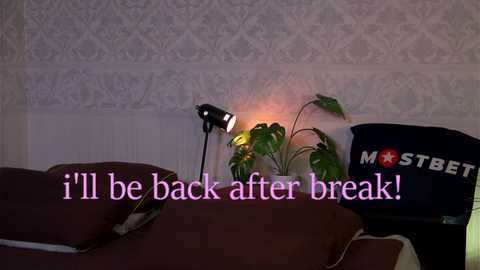 Media: A video of a dimly-lit bedroom featuring a maroon bed with a plush cover, a potted plant, and a \"MOSTBET\" baseball cap on a chair. The text reads, \"I'll be back after break!\