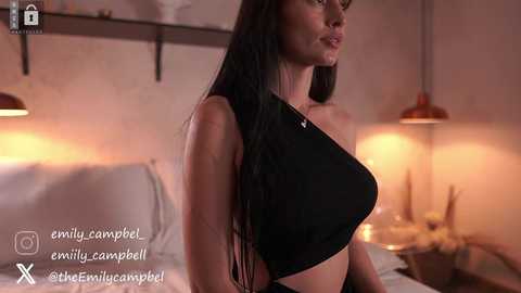 Media: Video of a fit woman with long black hair in a black crop top, standing in a dimly-lit bedroom with a bed, lamps, and a wooden shelf in the background.