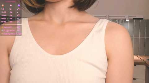 Media: Video of a woman with short brown hair, wearing a light beige ribbed tank top, showing her shoulders and upper chest. Background shows a blurred indoor setting with a window and a tiled wall.