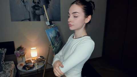 Media: A video of a young woman with pale skin, dark hair tied back, in a white sweater, standing in a dimly lit room with an easel, painting, and a lit candle on a table.