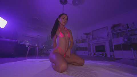 Media: Video of a slender, fit woman with long dark hair in a neon pink bikini, kneeling on a mat in a dimly lit, purple-lit room with shelves and kitchen items in the background.