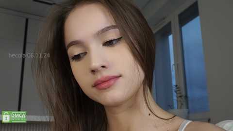 Media: Video of a young, light-skinned woman with straight, shoulder-length brown hair, wearing a white tank top, standing indoors near a window with a blurred blue sky background.