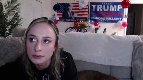 Media: Video of a blonde woman with a neutral expression sitting on a beige couch, with \"TRUMP\" banners and a vase of flowers in the background.