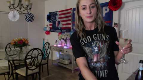 Media: A video of a blonde woman in a black \"God Guns and Guitars\" shirt, holding a champagne glass, standing in a decorated living room with American flags and party streamers.