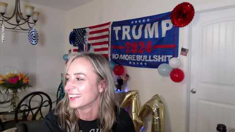 Media: Video of a smiling, fair-skinned, blonde-haired woman wearing a black shirt, sitting indoors with a \"TRUMP 2024 NO MORE BULLSHIT\" banner, red and blue balloons, and a gold foil elephant.