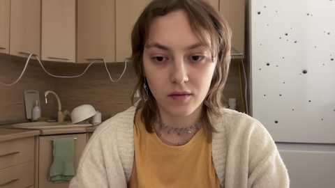 Media: Video of a young girl with shoulder-length brown hair, wearing a mustard-yellow shirt and beige cardigan, sitting in a kitchen with light wooden cabinets, a white stove, and a tiled backsplash.