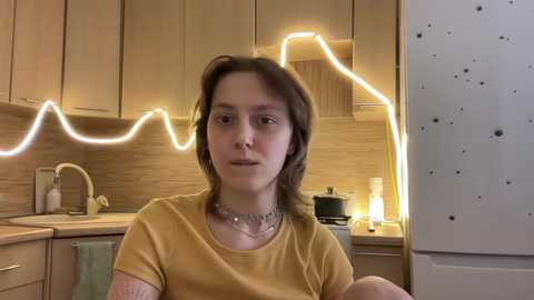 Media: Video of a young woman with light skin and brown hair, wearing a mustard-yellow t-shirt, in a modern kitchen with light wooden cabinets, a glowing neon light, and a microwave.