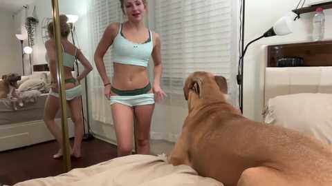 Media: Video of a muscular white woman in light blue sports bra and shorts, standing in a bedroom with white walls and beige bed, looking at a tan bulldog lying on the bed.