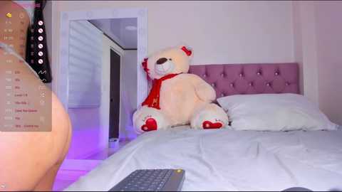 Media: A video of a plush teddy bear wearing a red scarf, sitting on a white bed with a purple tufted headboard in a softly lit bedroom. A remote control is visible on the bed.