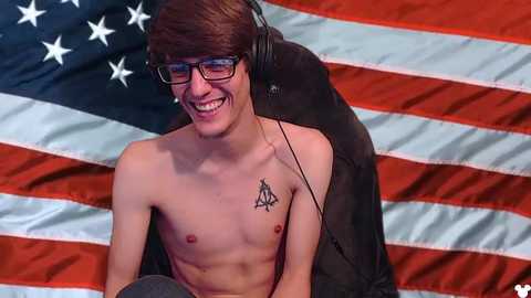 Media: Video of a shirtless, smiling young man with glasses, headphones, and a tattoo of a triangle on his chest. He sits against an American flag background.