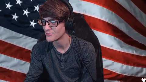 Media: Video of a young man with glasses, dark hair, wearing a gray shirt, headphones, sitting in a black chair in front of an American flag background.