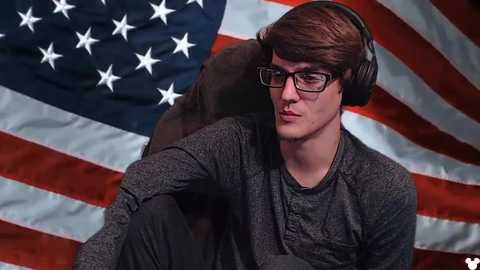 Media: Video of a young man with glasses, wearing a gray sweater and headphones, sitting against an American flag backdrop, appearing introspective.