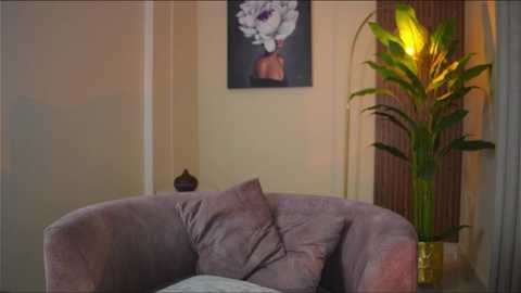 Media: A video of a cozy living room featuring a purple armchair with a decorative pillow, a tall, green potted plant with a yellow light, and a framed flower painting on a beige wall.