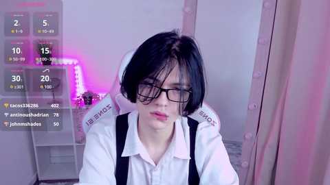 Media: Video of an East Asian woman with short black hair, glasses, and black suspenders, sitting in a room with a pink light and a \"JOY\" hoodie.