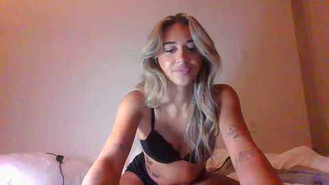 Media: Video of a smiling, light-skinned woman with long, wavy blonde hair, wearing black lingerie, sitting on a bed in a dimly-lit room with beige walls.