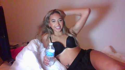 Media: Video of a smiling, light-skinned woman with long, wavy blonde hair, wearing a black bra and black shorts, holding a water bottle. She is lying on a white bed in a dimly lit room.