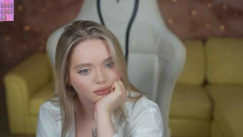 Media: Video of a blonde woman with fair skin, blue eyes, and a white shirt, seated on a yellow couch with a plush white chair behind her.