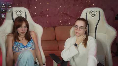 Media: Video of two women in gaming chairs, one with long brown hair in a blue bra, the other with glasses, white hoodie, and hand on her neck, in a dimly lit room with pink walls and a beige sofa.