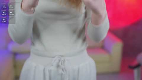 Media: A video of a fair-skinned woman in a light gray, long-sleeved dress, holding her hands up, blurred background with pink and purple hues.